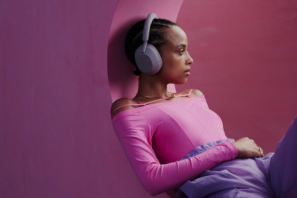 A promotional shot of a girl leaning against a pink wall wearing her WH-1000XM5 wireless headphones.